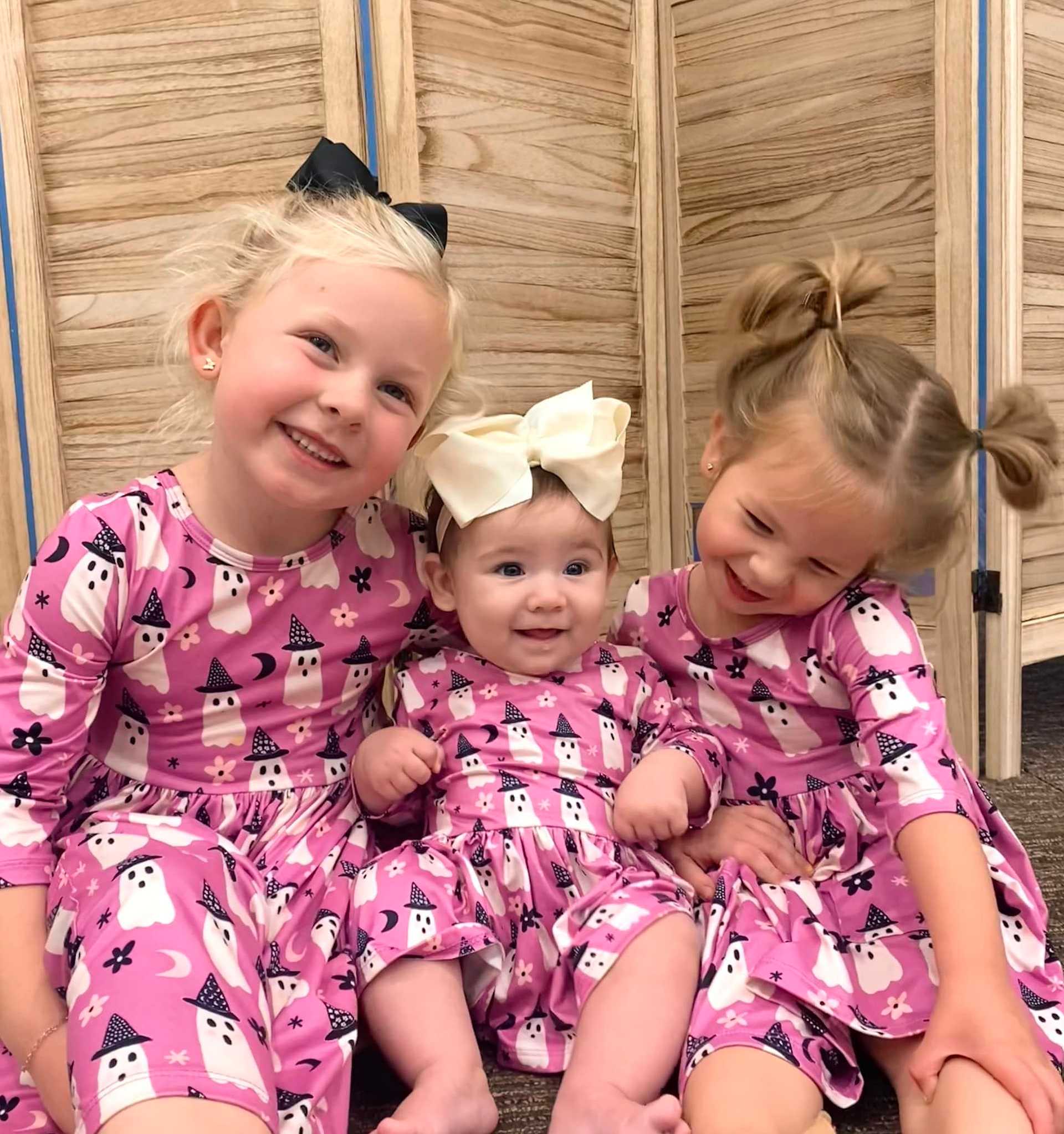 Three young girls in matching pink dresses with ghost prints, sitting and smiling together.