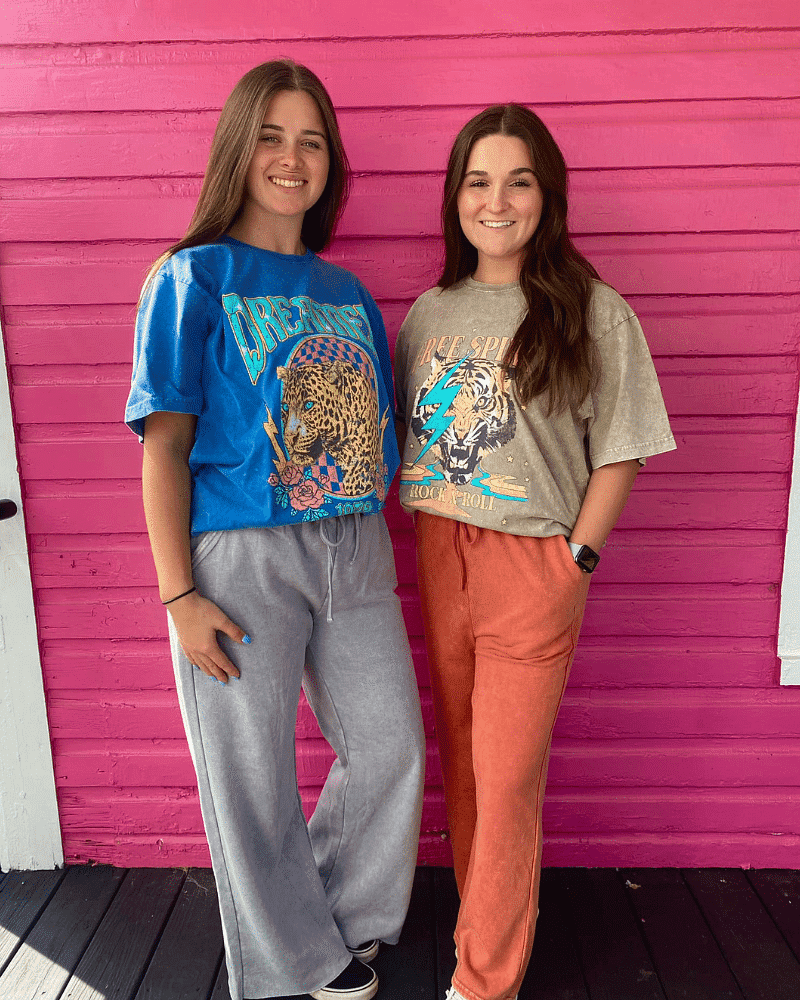 Two women standing and smiling, wearing colorful graphic T-shirts and casual pants, against pink wall.